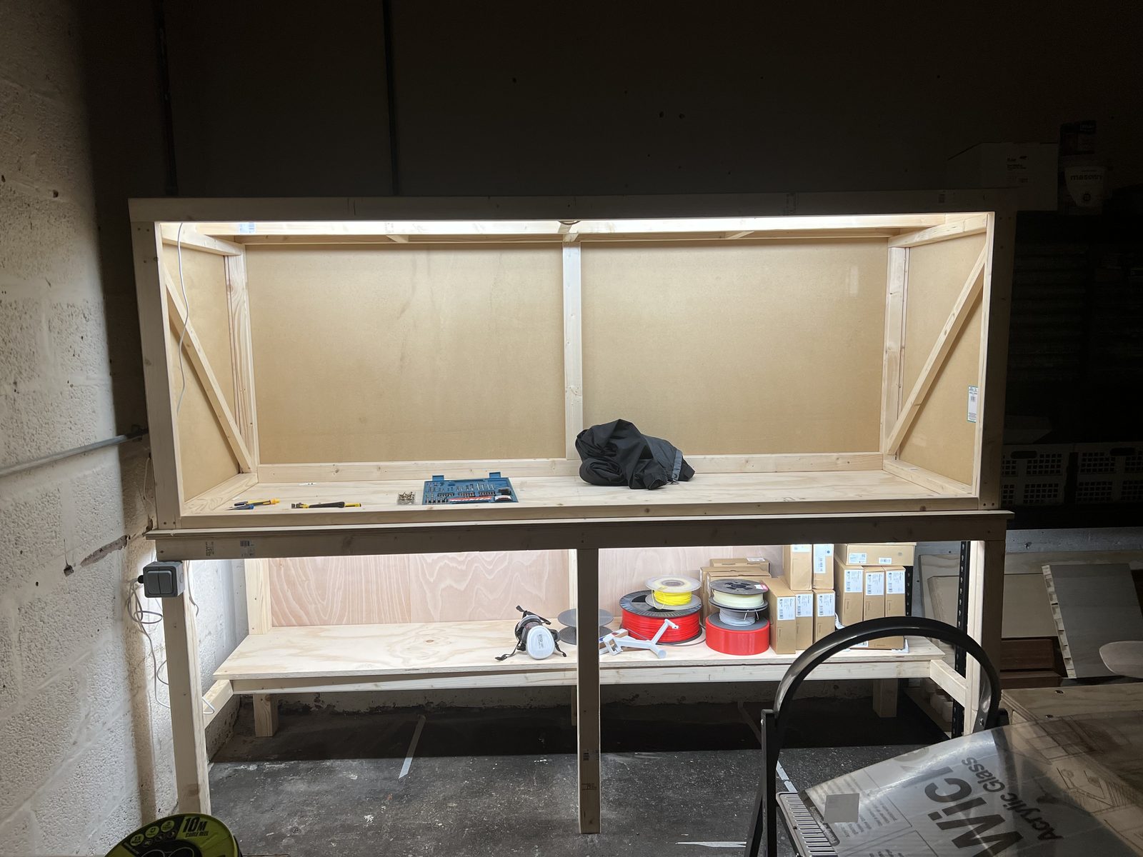 A newly built timber workbench with a lit upper cabinet, filament spools and boxes stacked on the shelf below.