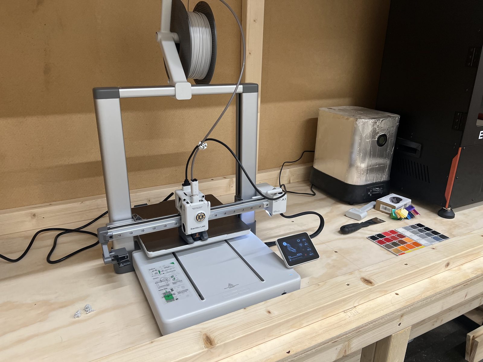 A Bambu Lab A1 3D printer on a workbench next to a foil-wrapped filament dryer and a resin printer.