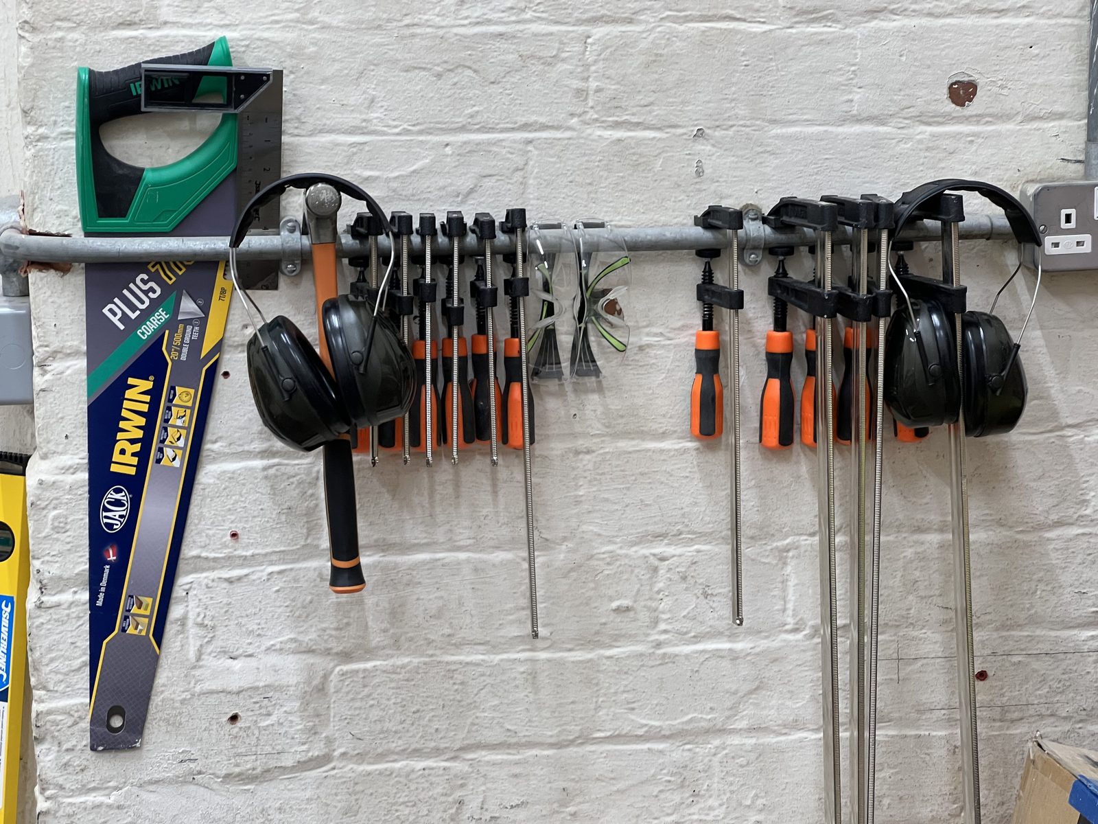 A tool rail on a white brick wall holding saws, clamps, screwdrivers, safety glasses and ear defenders.