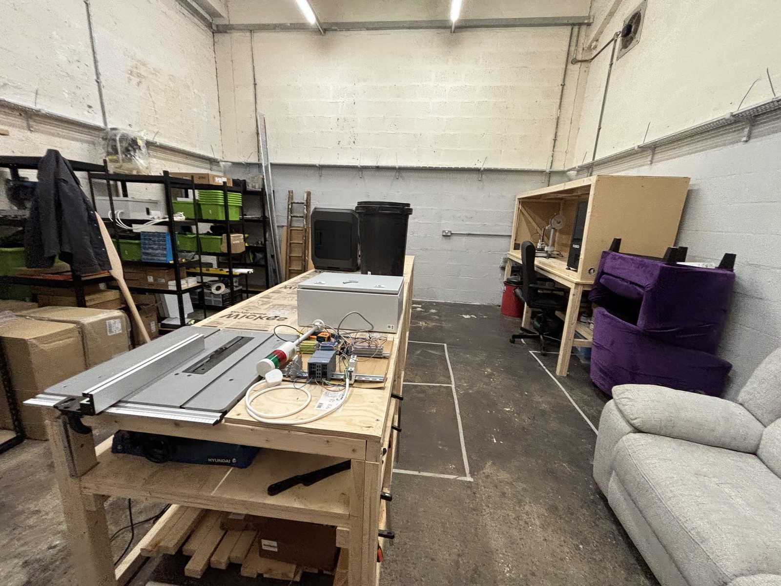 Inside the unit: a large workbench with a table saw down the middle, shelving with labelled crates, a bench with a mounted enclosure, and a sofa along the wall.