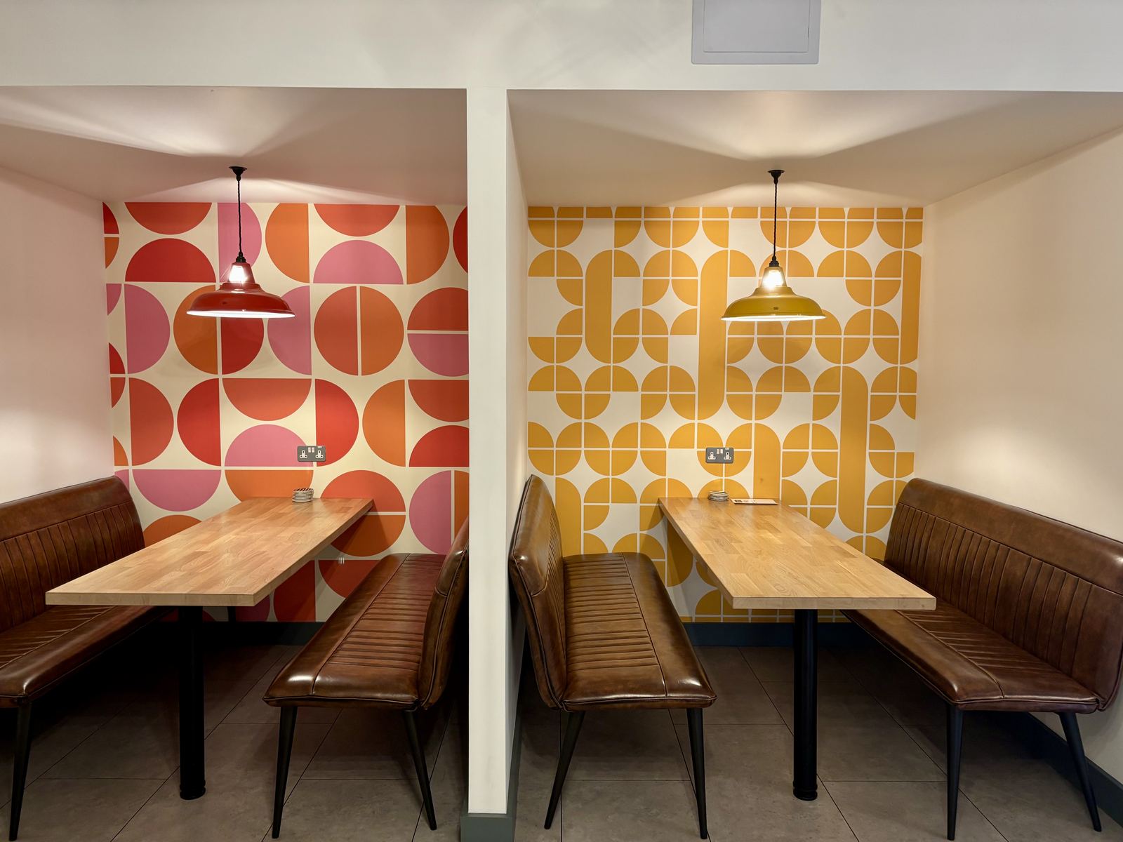 Two booths with leather bench seating and wooden tables, each under a pendant light, against bold retro geometric wallpaper in red and mustard.